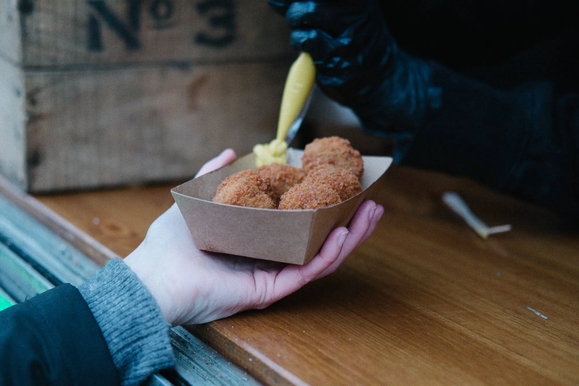 Hand houdt een bakje bitterballen vast terwijl er mosterd op wordt gespoten