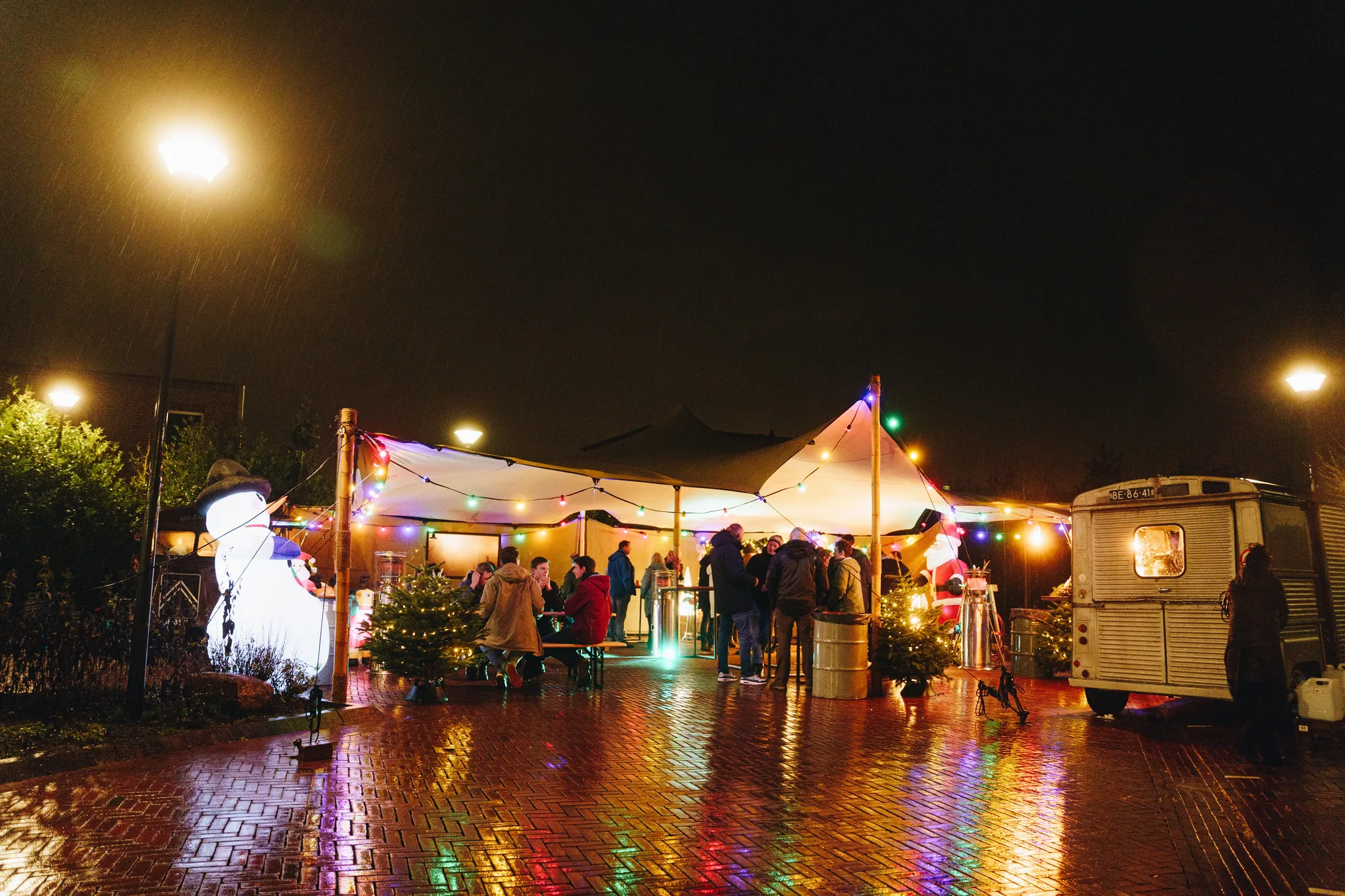 Verlichte stretchtent en foodtruck weerspiegeld in nat wegdek tijdens een winterfeest