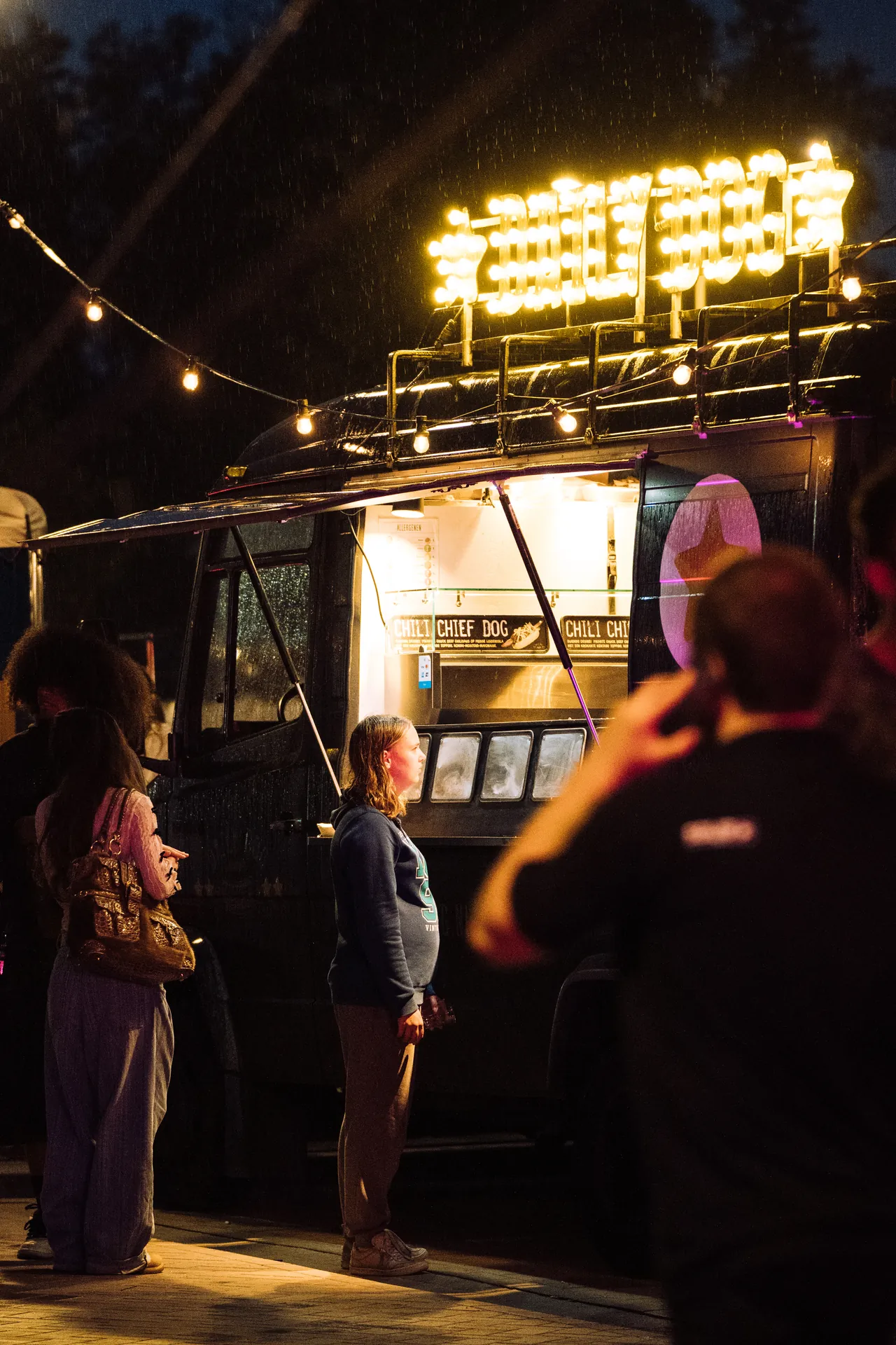Gasten wachten bij de verlichte Daily Dog foodtruck in de avond